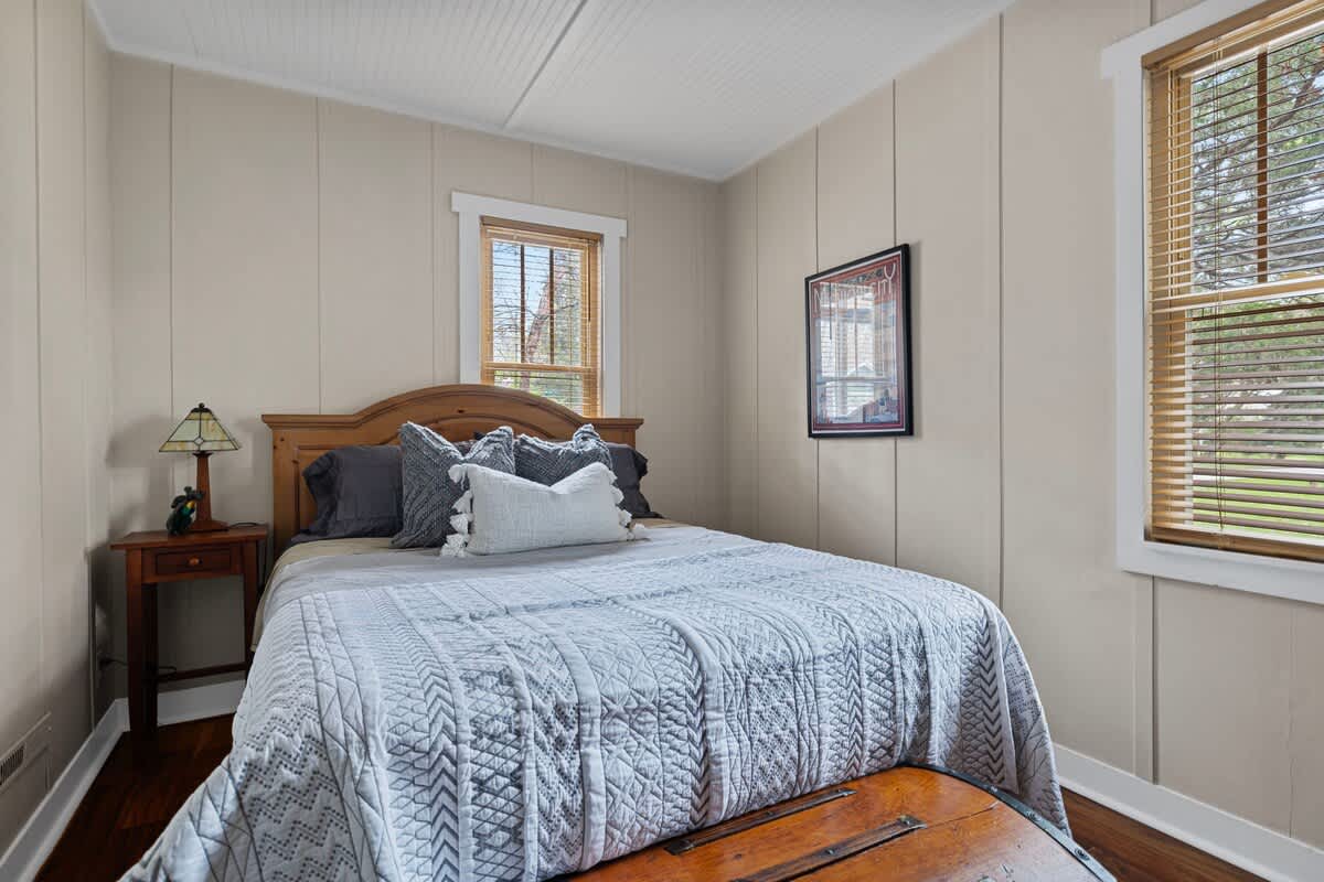 Cozy bedroom featuring a wooden headboard on a bed with a light gray quilt, a small wooden nightstand with a lamp, and two windows with wooden blinds, showcasing paneled walls.