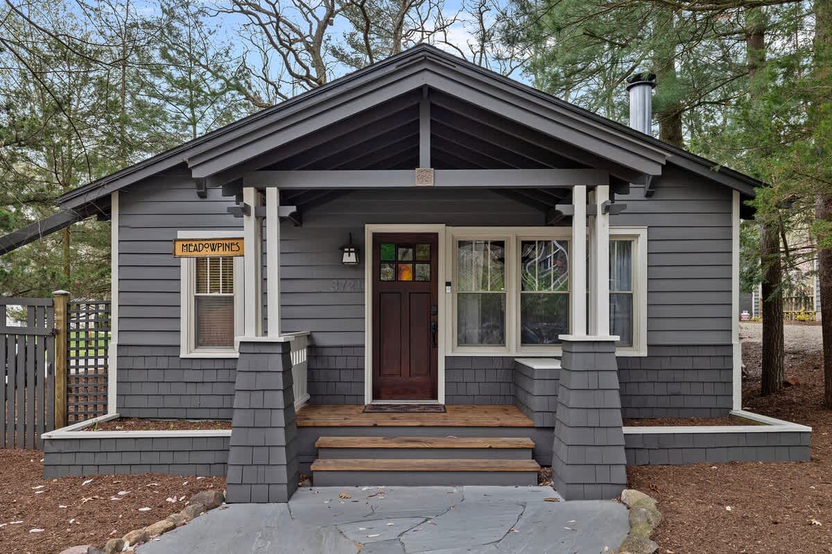 The charming front facade of a dark gray cottage-style home, featuring white trim, a covered porch with sturdy white pillars, and a dark wood front door with stained glass accents. The lower half of the house and porch pillars are clad in dark gray shingle siding, with a 'MEADOWPINES' sign visible.