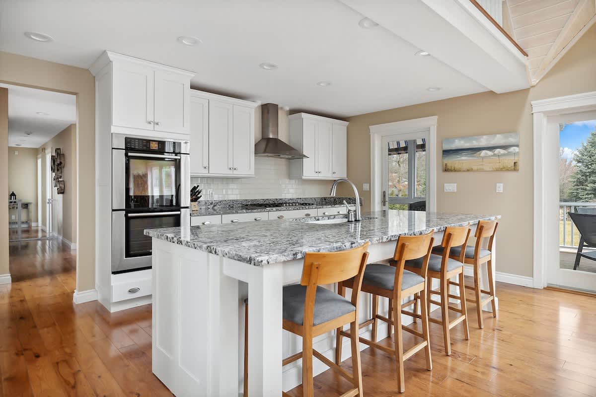 A modern kitchen boasts white shaker cabinets, granite countertops, and stainless steel appliances, including a double oven. A large island with bar seating for four overlooks the open living space.