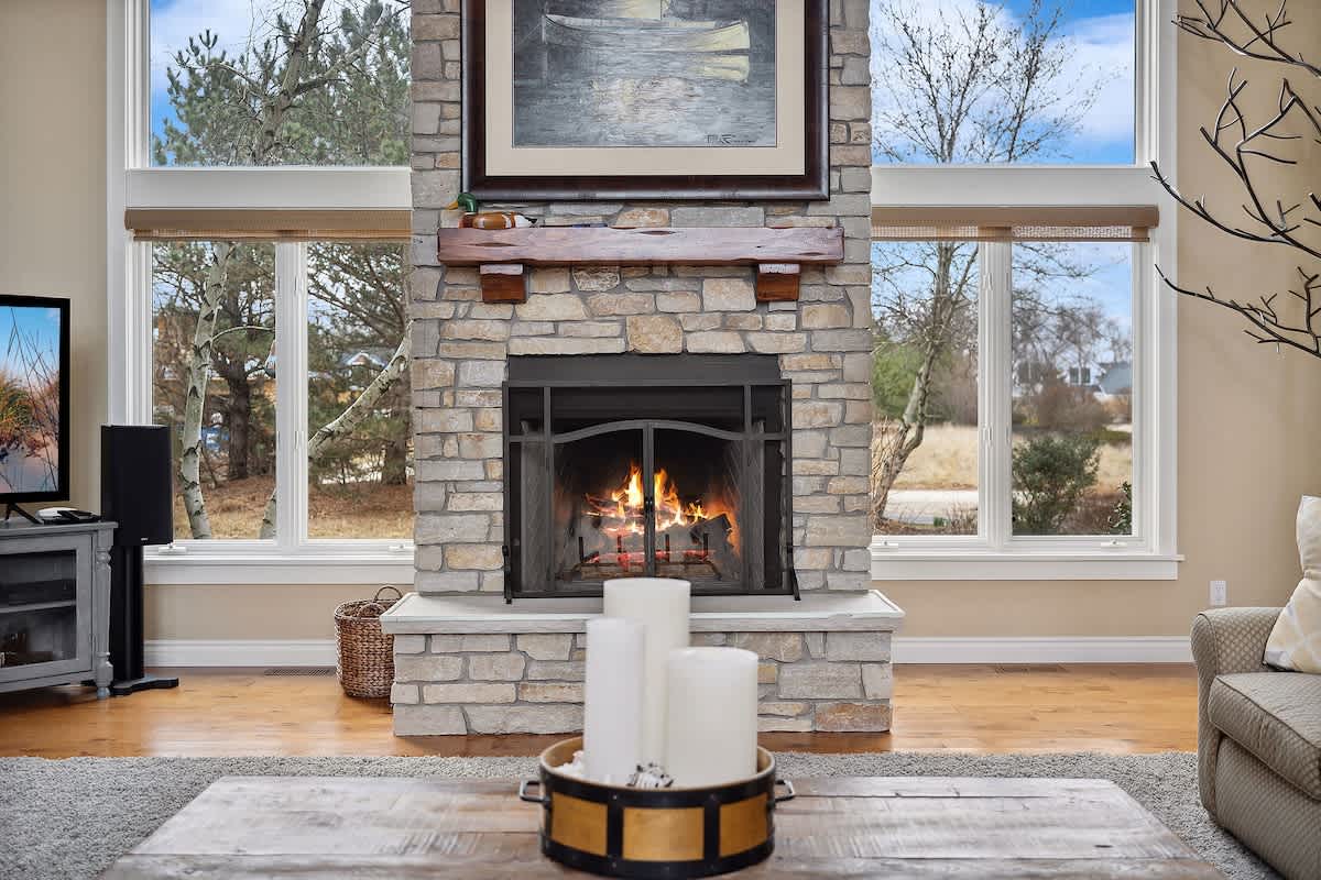 A cozy living room centers around a floor-to-ceiling stone fireplace with a wooden mantel, flanked by large windows that offer views of the outdoors, with a television and comfortable seating visible.
