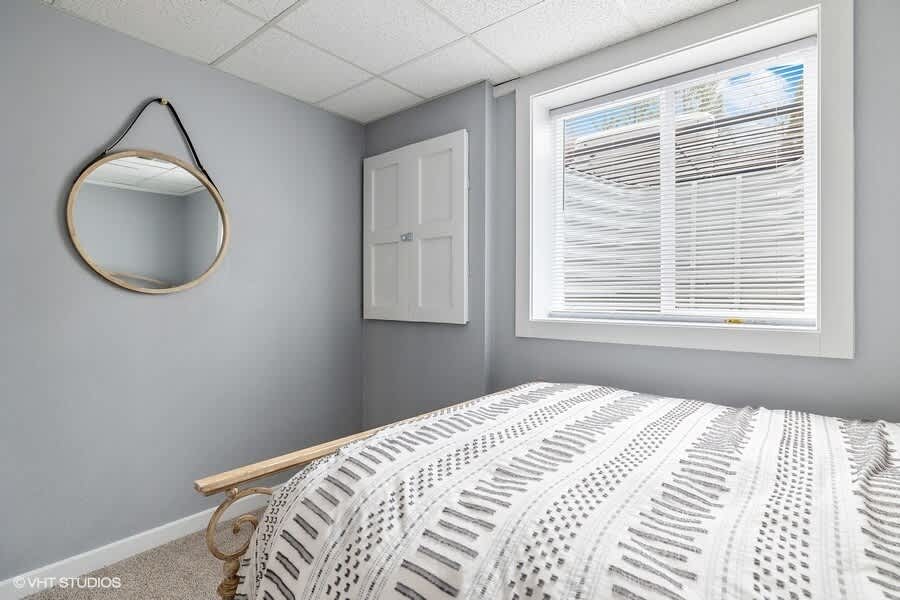 A different angle of the cozy bedroom, showing a round wooden mirror on the light gray wall, the metal-frame bed with patterned bedding, and a window with white blinds.