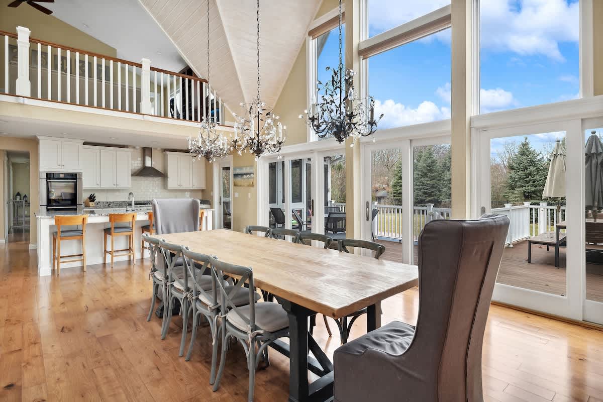 A long, rustic wooden dining table with grey chairs sits beneath three ornate chandeliers, facing a wall of tall windows and glass doors that open to an outdoor deck, with a modern white kitchen visible in the background.
