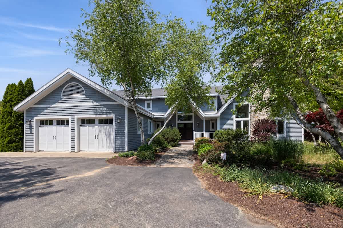 The front exterior of a light blue house with white trim and a gray roof, featuring a two-car garage with white doors and a stone facade accent. A paved driveway leads to a walkway surrounded by mature trees and landscaping.