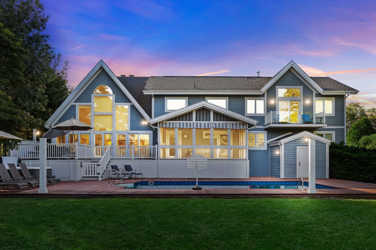 The impressive rear exterior of a large blue house with multiple gables and large windows, illuminated at dusk. A spacious wooden deck surrounds a rectangular swimming pool, with a grassy yard and mature trees in the foreground.