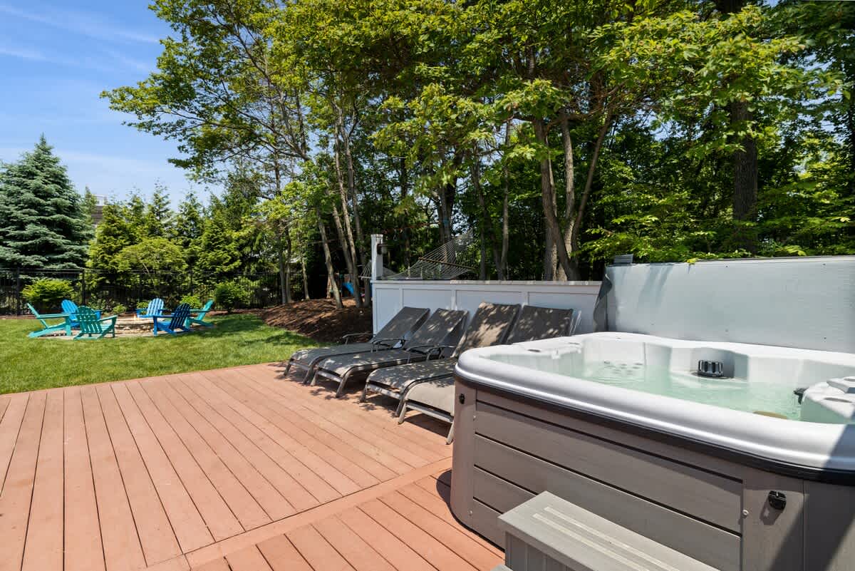 A modern grey hot tub is positioned on a wide wooden deck, accompanied by several dark lounge chairs, with a lush green lawn and dense trees providing a natural backdrop under a bright sky.