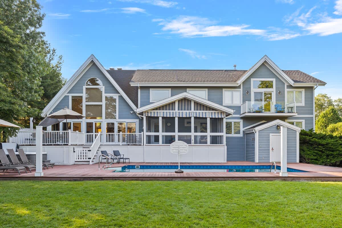 The expansive back of a multi-gabled blue house features a large deck with an in-ground swimming pool, a screened-in porch, and numerous windows, all overlooking a green lawn under a bright blue sky.