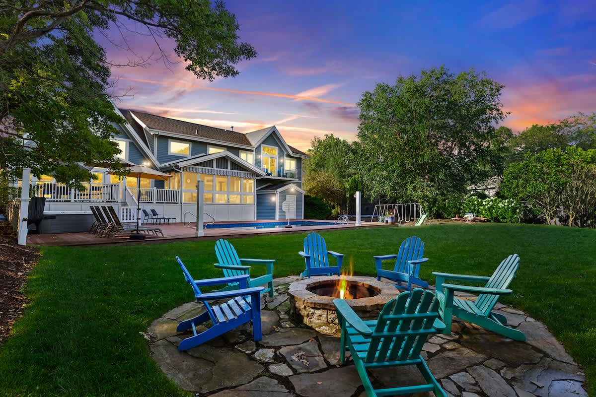 A cozy outdoor fire pit area with a stone base and several blue and teal Adirondack chairs arranged around it, set on a flagstone patio. The lush green lawn extends towards the large blue house with a pool deck, all under a vibrant sunset sky.