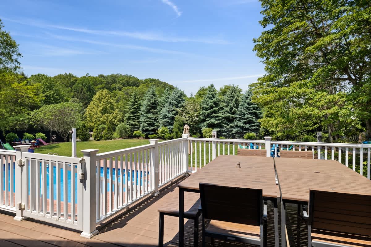 A spacious wooden deck with a large outdoor dining table and chairs, enclosed by a white railing, offering a view of the swimming pool and the extensive green backyard.