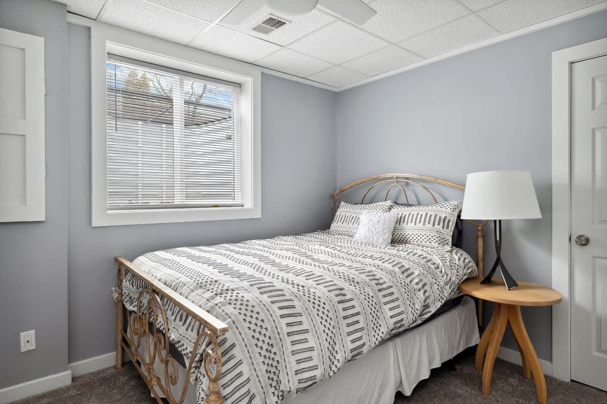 A cozy bedroom featuring light gray walls, a metal-frame bed with a patterned duvet, a wooden nightstand, and a window with blinds, all under a white acoustic tile ceiling.