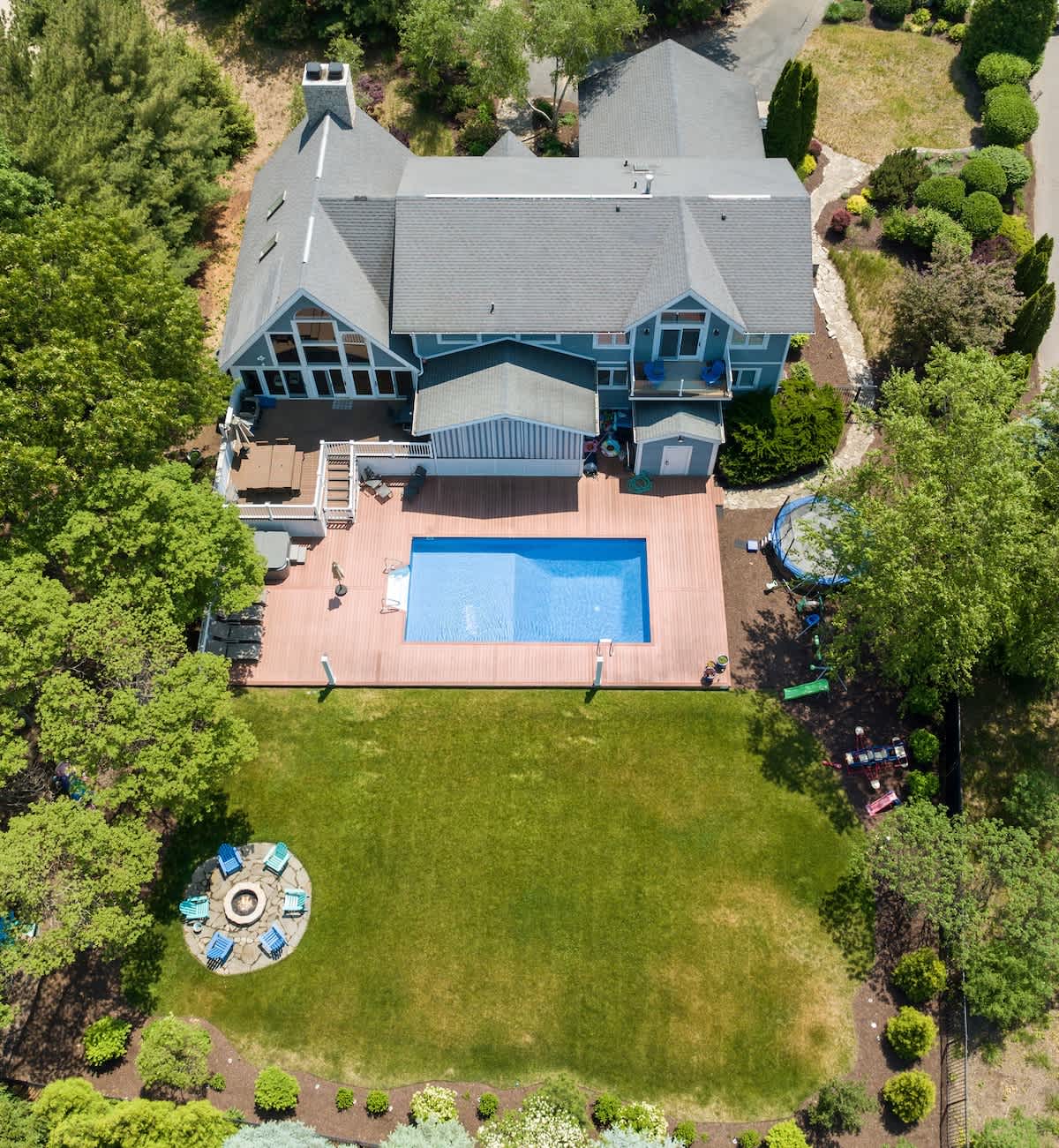 Overhead aerial view showcasing a large blue house with a gray roof, a rectangular swimming pool surrounded by a wooden deck, and an expansive green lawn with a stone fire pit area. The property is surrounded by mature trees and landscaping.