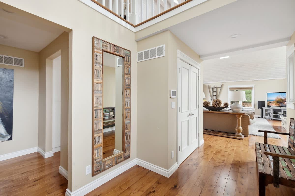 A bright foyer with light beige walls and hardwood floors features a large, rustic-framed mirror and a wooden bench, leading into an open-concept living area with high ceilings.