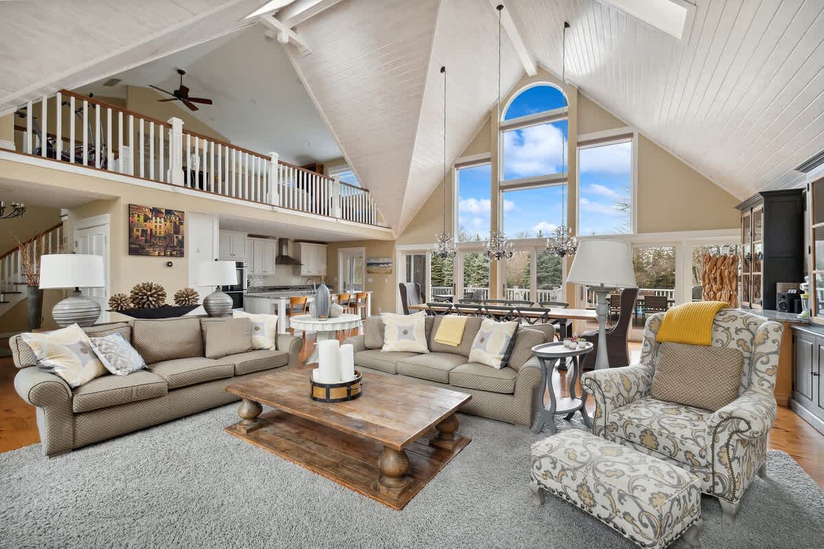 A spacious living room features two large sofas and an armchair arranged around a rustic wooden coffee table on a grey rug, with vaulted ceilings, large windows, and an open-concept view to the dining area and kitchen.