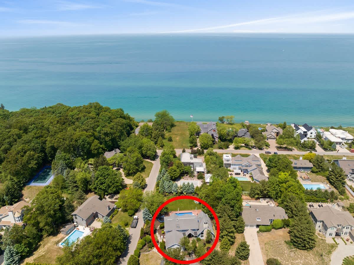 Expansive aerial view showing the property, highlighted by a red circle, situated amidst other homes and lush greenery, with a large body of water (lake or ocean) in the background under a clear sky.