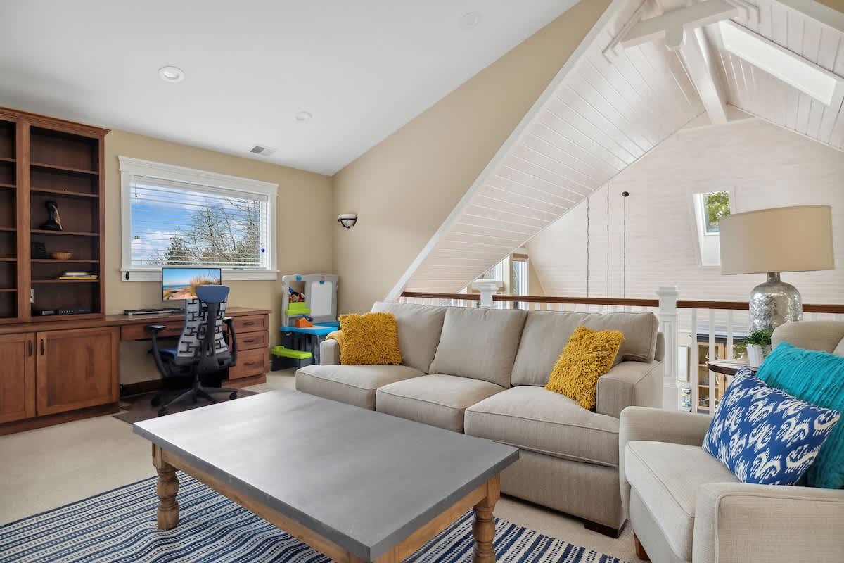 A bright loft living area features a comfortable sofa and armchair, a large coffee table, and a built-in wooden desk with shelving. The space overlooks the lower level and includes a window with outdoor views.