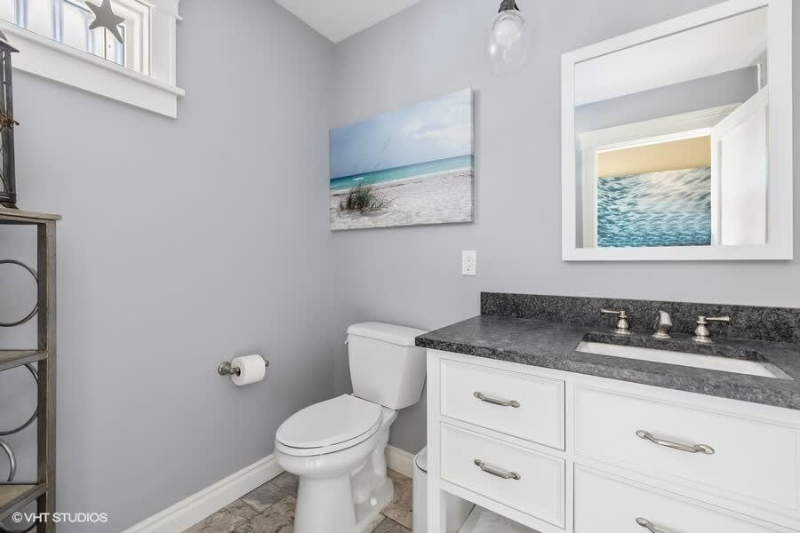 A modern bathroom with light gray walls, a white vanity with a dark granite countertop and single sink, a toilet, and a framed beach artwork above the toilet.