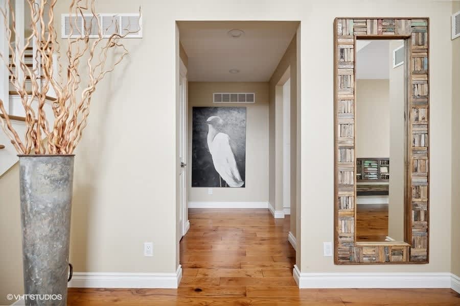 A bright foyer features light beige walls, hardwood floors, and a large decorative mirror with a rustic frame. A tall vase with dried branches stands next to a white staircase, leading into a hallway with a large bird painting.