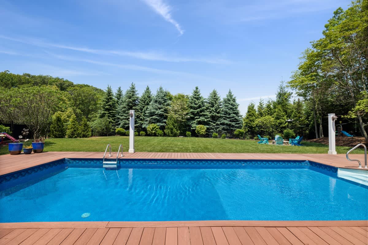 A wide shot of the outdoor swimming pool and expansive green lawn, bordered by a dense line of evergreen trees under a clear blue sky.