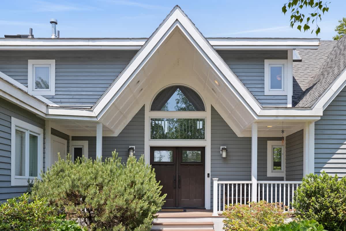 The front entrance of a grey-sided house features a prominent gabled portico with an arched window above dark double doors, flanked by white railings and lush green bushes.