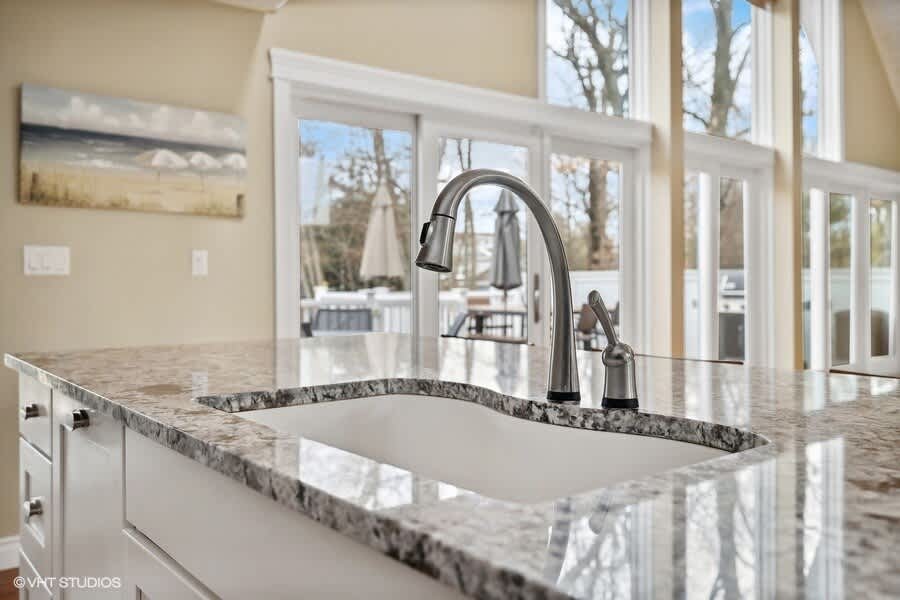 A close-up of the kitchen island highlights the polished granite countertop and a modern gooseneck faucet over a white undermount sink. Large windows in the background offer views of the outdoor deck and greenery.