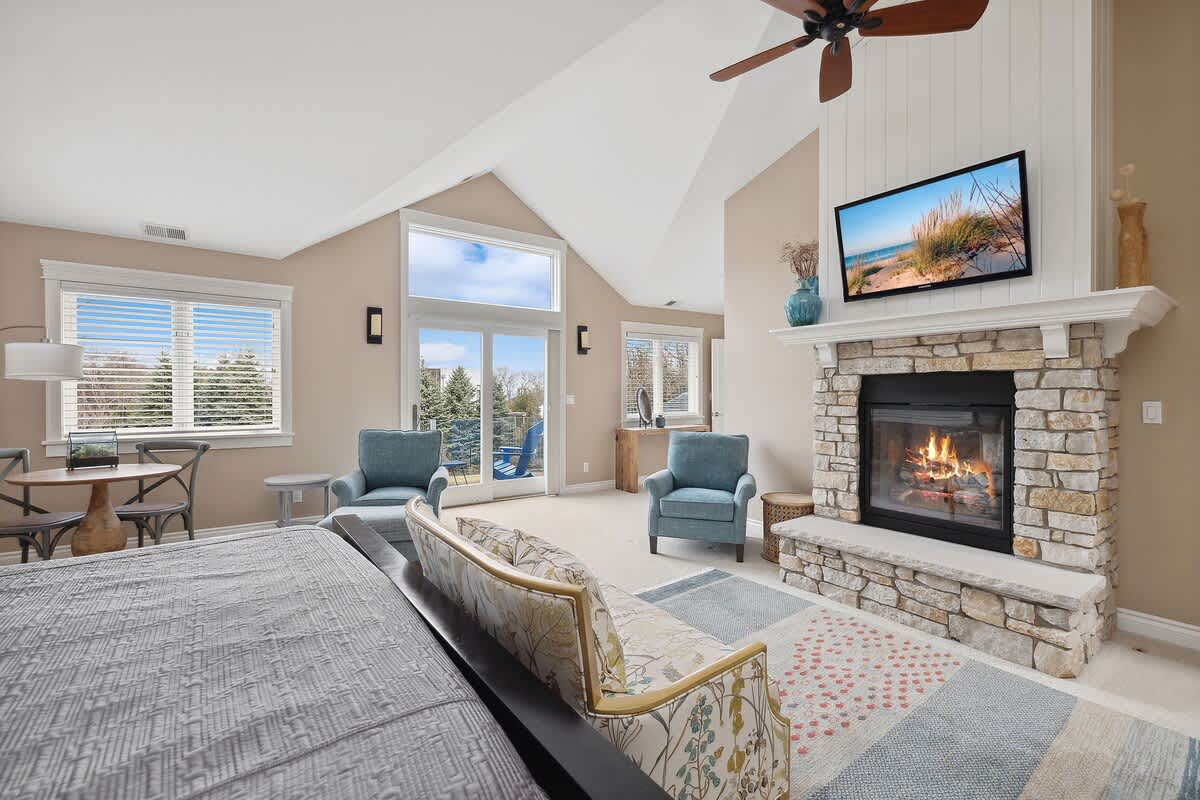 Master bedroom featuring a stone fireplace with a white mantel, a wall-mounted TV, and a seating area with two blue armchairs, all under a high vaulted ceiling with a ceiling fan.