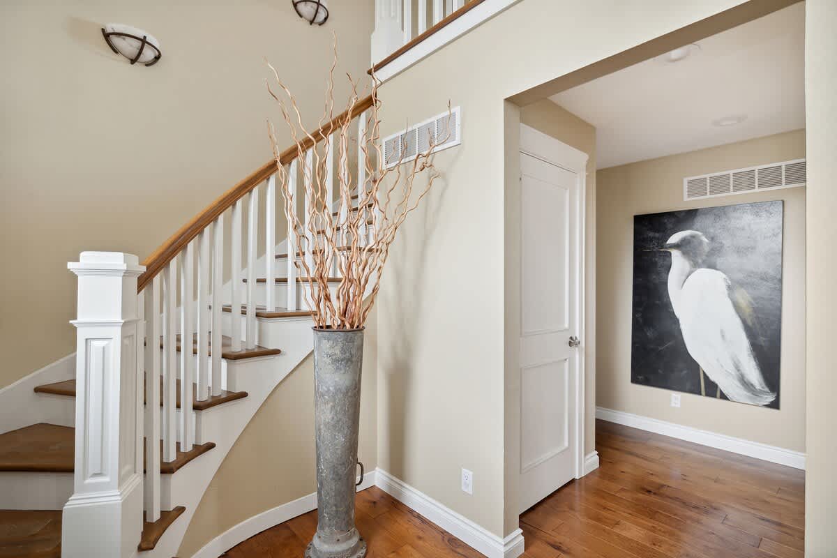 A welcoming foyer showcases a white banister staircase with wooden handrails and hardwood floors. Light beige walls lead into a hallway adorned with a striking black and white painting of a bird.