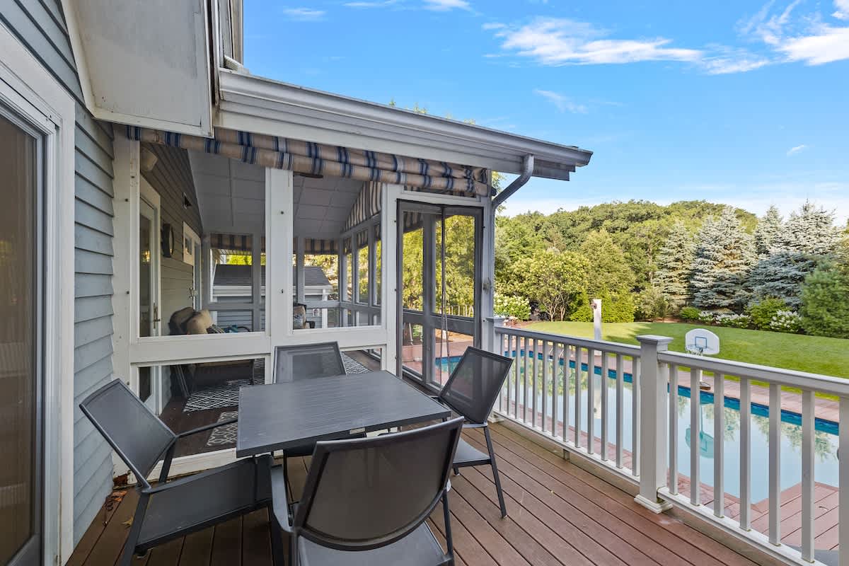A multi-level wooden deck featuring an outdoor dining set with a black table and chairs, overlooking the swimming pool and a lush green backyard, with a screened-in porch visible on the left.