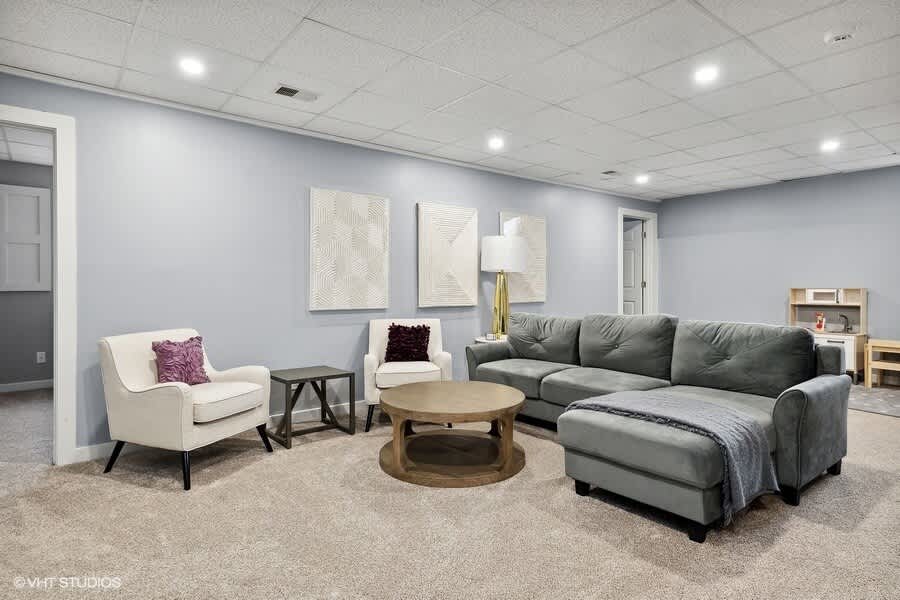 Spacious living room with light blue walls, a large gray sectional sofa, two cream armchairs, a round wooden coffee table, and recessed lighting, featuring a children's play kitchen in the background.