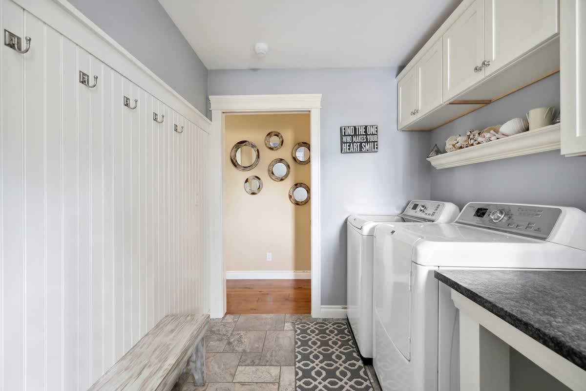 A bright laundry room featuring a white washer and dryer, dark speckled countertop, white upper cabinets, and a white beadboard wall with hooks and a light wood bench. A patterned rug covers part of the tiled floor, and a doorway leads to a room with multiple round mirrors.