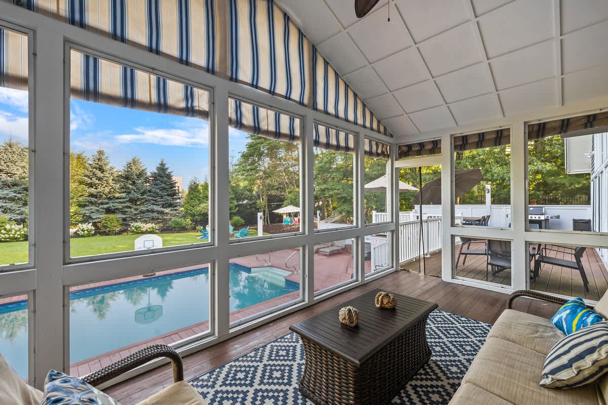 This sun-filled screened porch provides a comfortable seating area with wicker furniture and a dark patterned rug. It offers expansive views of the outdoor swimming pool and an adjacent deck with additional seating.