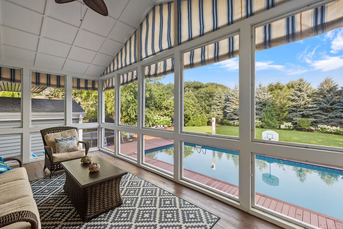 A cozy screened-in porch features wicker furniture with comfortable cushions and a patterned outdoor rug. Large windows offer panoramic views of the private swimming pool and lush backyard.