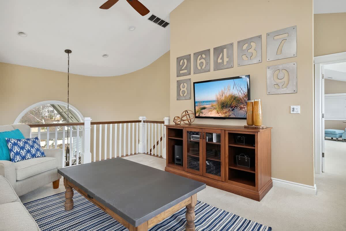 This cozy loft living space features a flat-screen TV on a wooden media console, surrounded by unique metal number wall art. A comfortable sofa and armchair are arranged on a striped rug, with a railing overlooking the floor below.