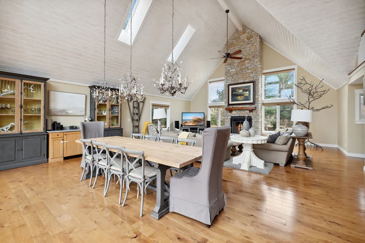 An expansive open-concept living and dining area features vaulted ceilings with skylights and multiple chandeliers. Hardwood floors extend throughout, leading to a stone fireplace and large windows overlooking the outdoors.