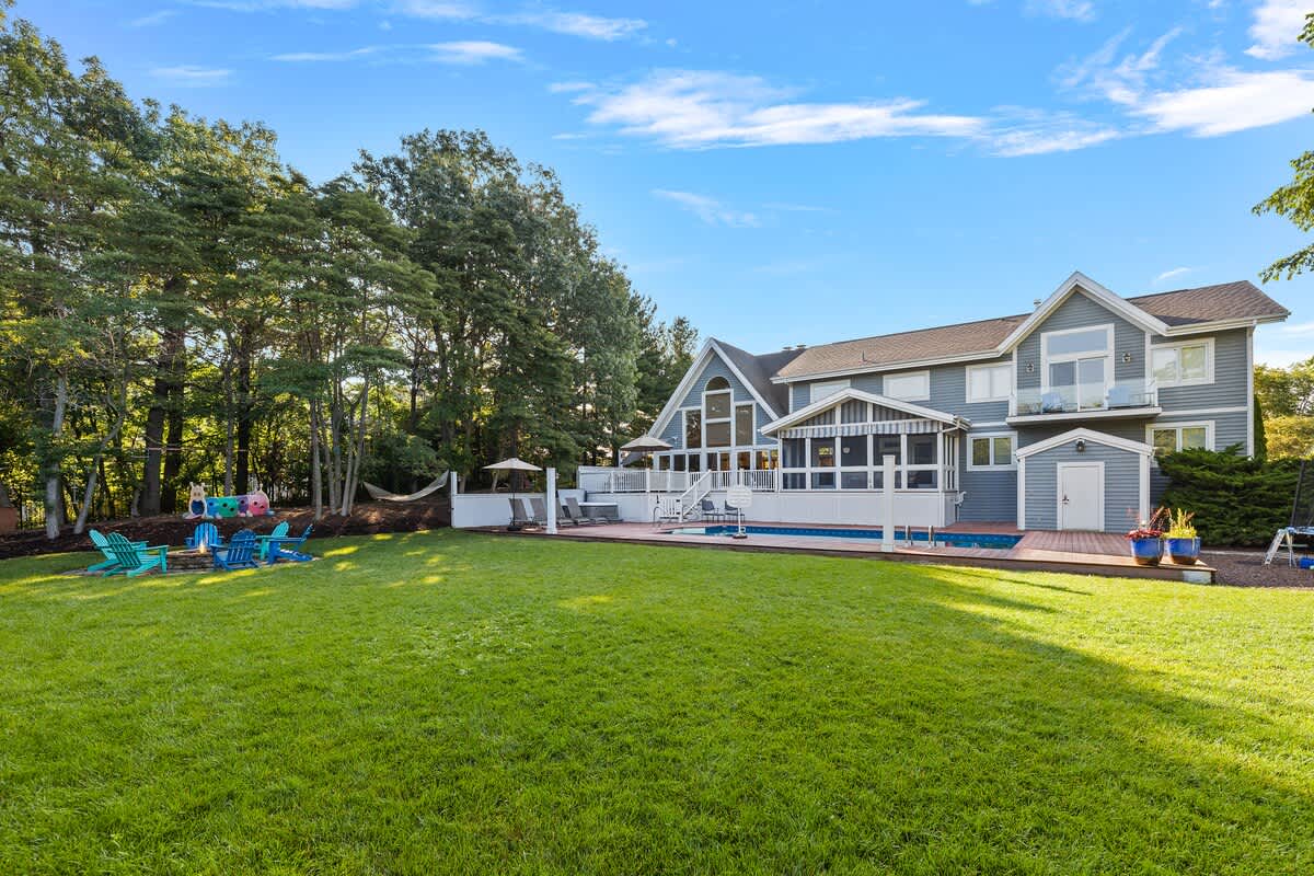 A vast green lawn stretches out towards a large blue house with a pool and deck, featuring a fire pit area with colorful Adirondack chairs and a children's play area nestled among mature trees, all under a clear blue sky.