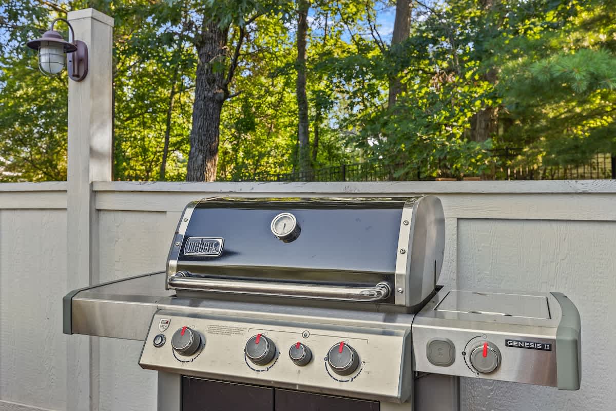 A modern stainless steel Weber Genesis II gas grill with a black lid, positioned on a wooden deck or patio, surrounded by a light-colored fence and lush green trees in the background.