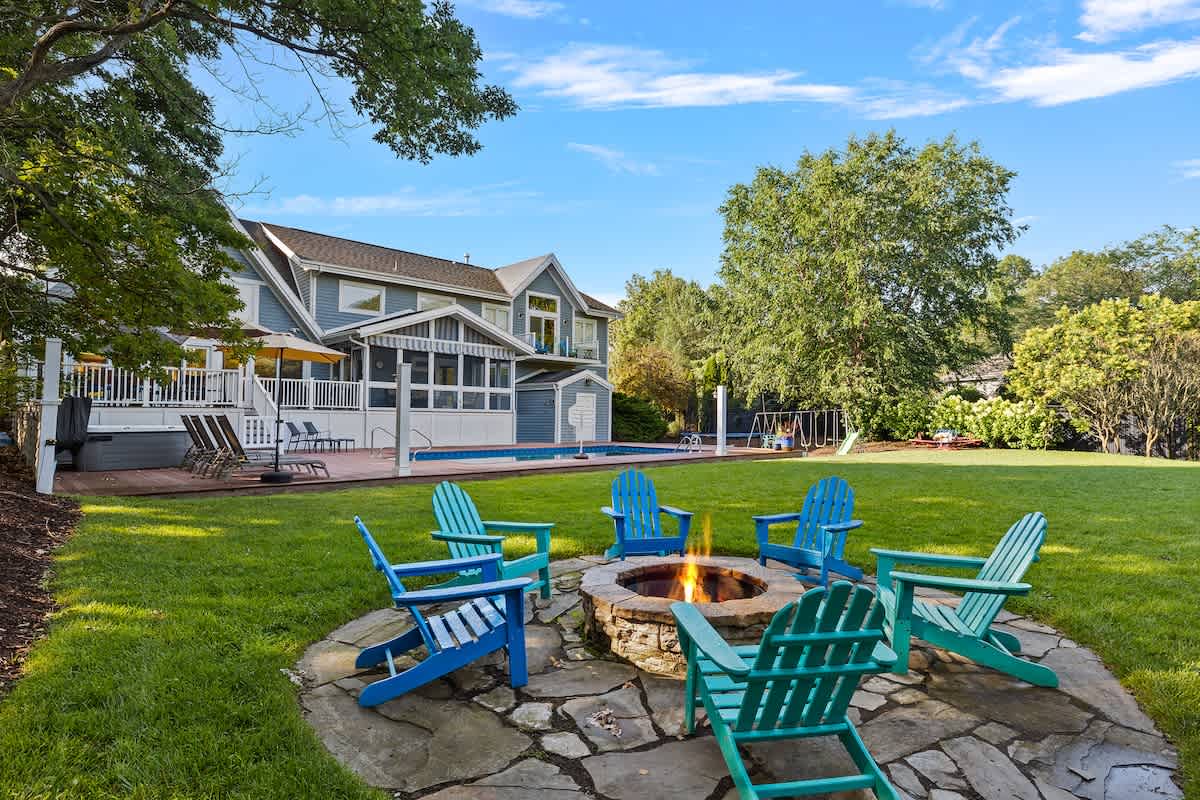 A stone fire pit with a roaring fire is surrounded by blue and teal Adirondack chairs on a flagstone patio, set on a lush green lawn with a large house and pool in the background under a blue sky.
