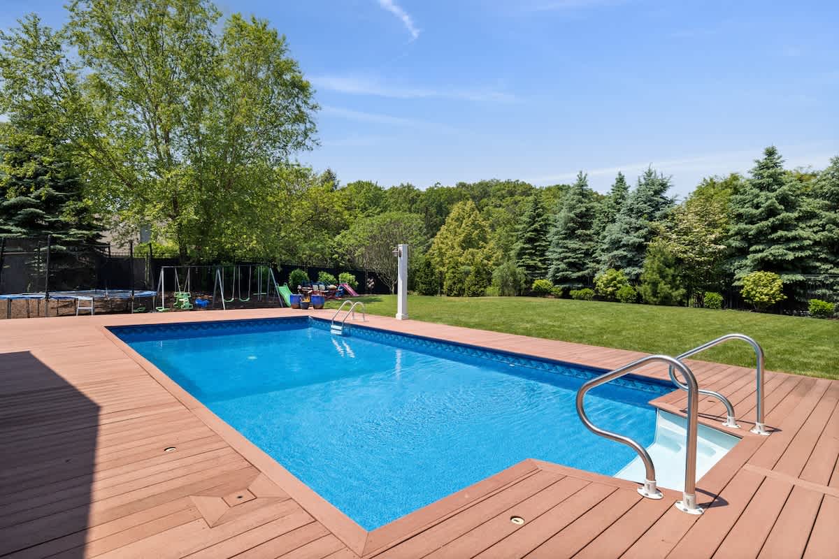 A large rectangular outdoor swimming pool with bright blue water, surrounded by a spacious wooden deck, set in a lush green backyard with mature trees.