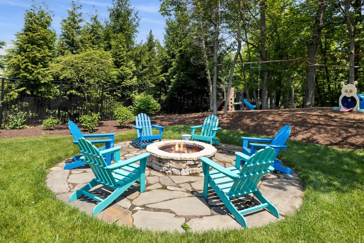 A circular stone fire pit surrounded by six colorful Adirondack chairs (blue and teal) on a flagstone patio, set within a grassy area bordered by trees and a children's play structure.
