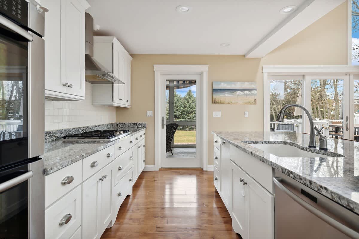 This bright kitchen features white cabinetry, speckled granite countertops, and a white subway tile backsplash. A stainless steel range hood and cooktop are visible, with a glass door leading to an outdoor area.