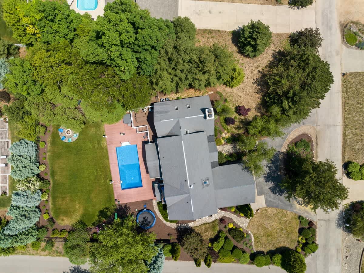 High aerial view of a large house with a dark gray roof, surrounded by lush green trees and landscaping. A rectangular swimming pool with a wooden deck is visible in the backyard, along with a grassy lawn and a trampoline.