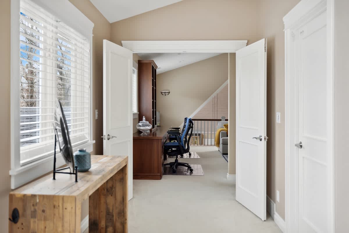 Bright office space with light beige walls, a wooden desk, and a black office chair, featuring white double doors opening to a landing with a built-in bookshelf and a window with blinds.