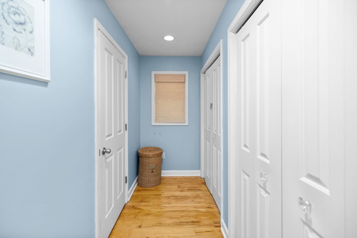 Bright hallway with light blue walls and warm hardwood floors, featuring multiple white doors leading to other rooms or closets. A small window with a blind and a wicker basket are visible at the end of the hall.