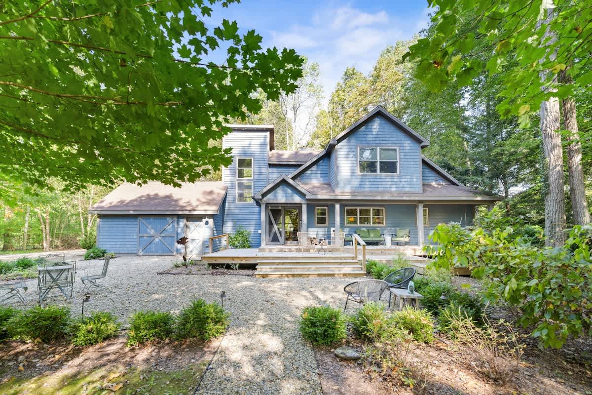 The back of a blue-sided house with multiple gables and a large wooden deck is surrounded by mature green trees, featuring a gravel path and outdoor seating areas.