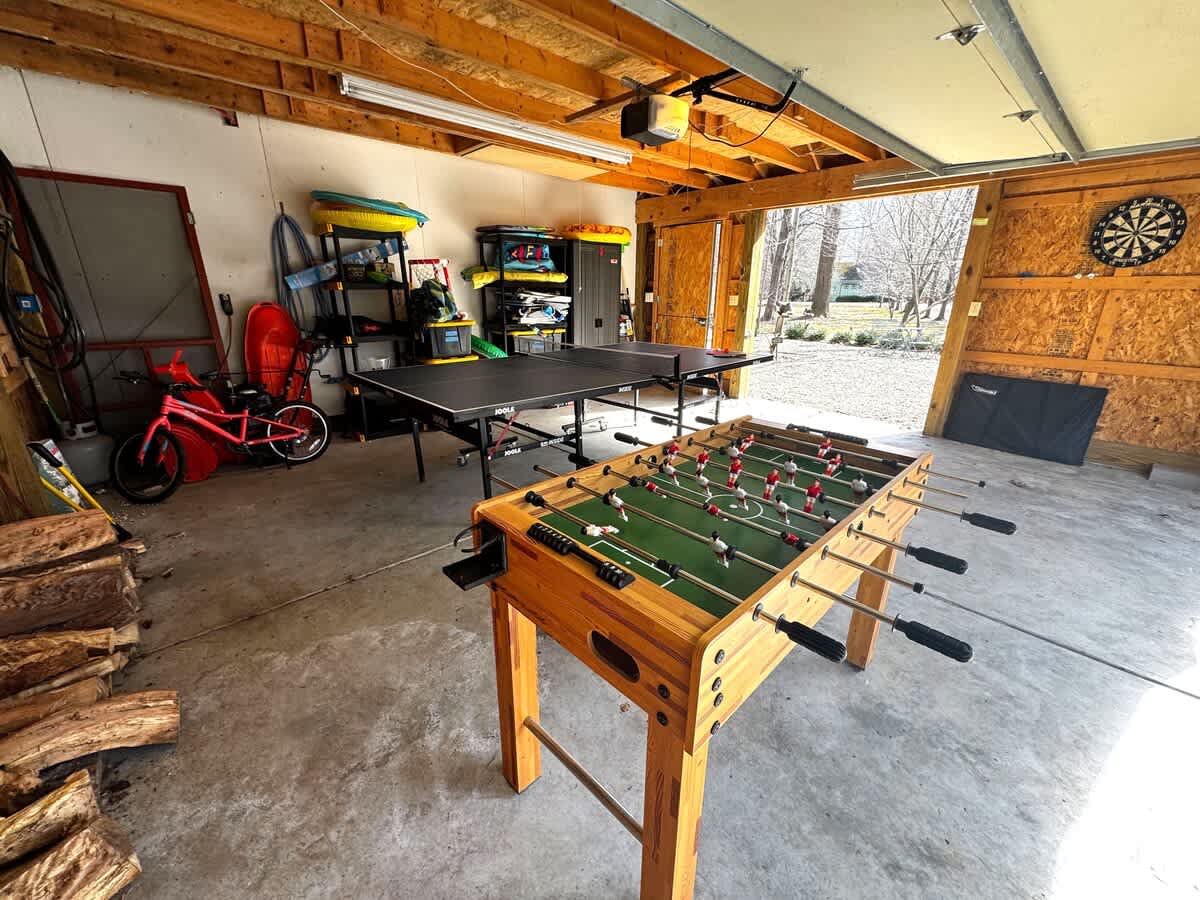 Spacious garage converted into a game area, featuring a black ping-pong table and a wooden foosball table on a concrete floor. Various storage shelves, a red bicycle, and a dartboard are visible, with the garage door open to an outdoor view.