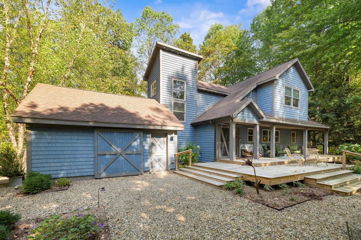 The front exterior of a blue-sided house with a large wooden deck and a gravel driveway, featuring a barn-style garage door and surrounded by lush green trees.