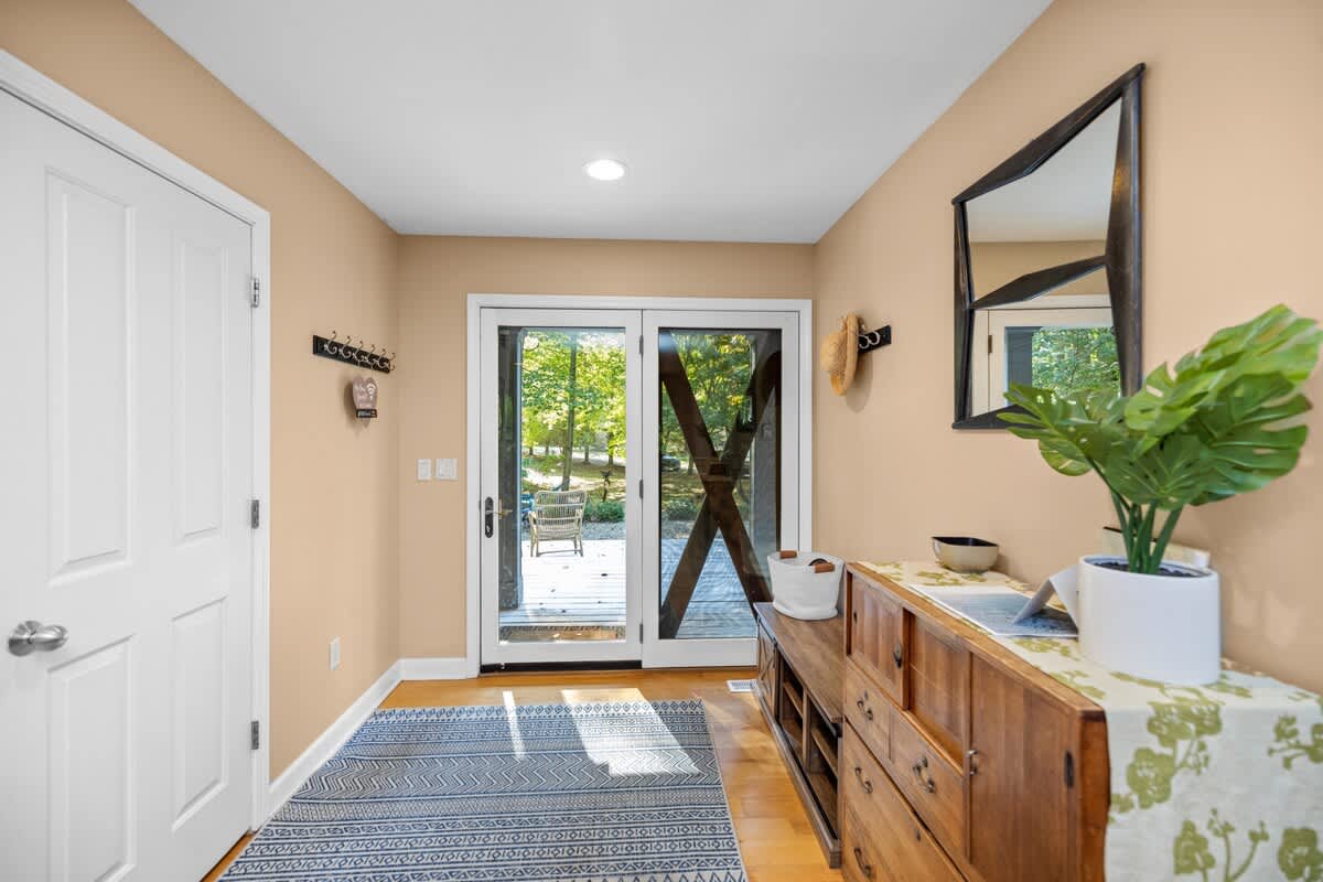 A bright foyer with light peach walls and hardwood floors features a wooden console table with a plant, a large dark-framed mirror, and sliding glass doors leading to the exterior.