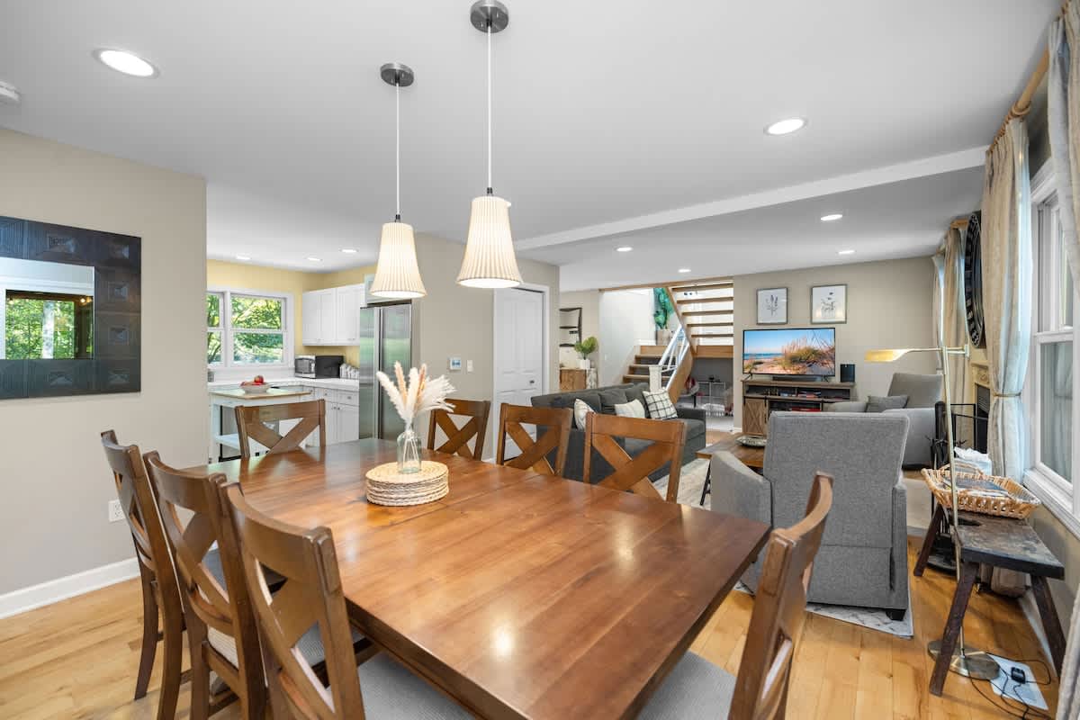 A dining area with a large wooden table and eight chairs is brightly lit by pendant lights, flowing into a living room with a fireplace and a kitchen visible in the background.