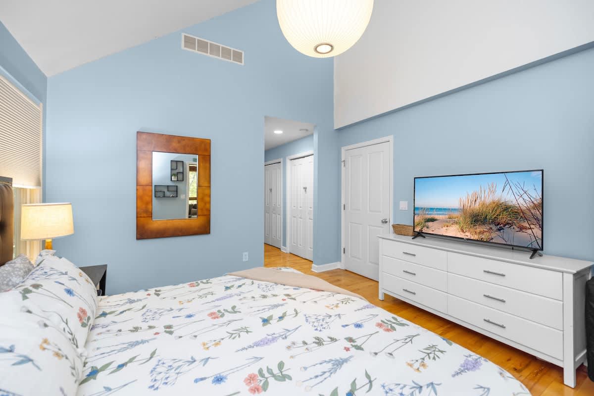 Spacious bedroom with light blue walls and a vaulted white ceiling. A bed with a floral duvet is positioned next to a nightstand with a lamp, and a large flat-screen TV sits on a white dresser opposite the bed.