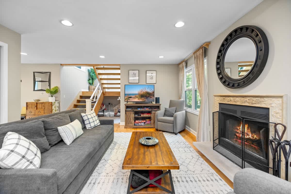 A spacious living room with light hardwood floors and neutral walls features a large gray sectional sofa, a wooden coffee table, a TV on a console, and a stone fireplace, with a staircase visible in the background.