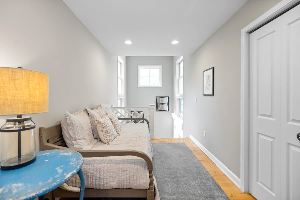 Bright loft area with light gray walls and hardwood floors, featuring a daybed with pillows and a round blue side table. A railing overlooks a lower level, and a window provides natural light.