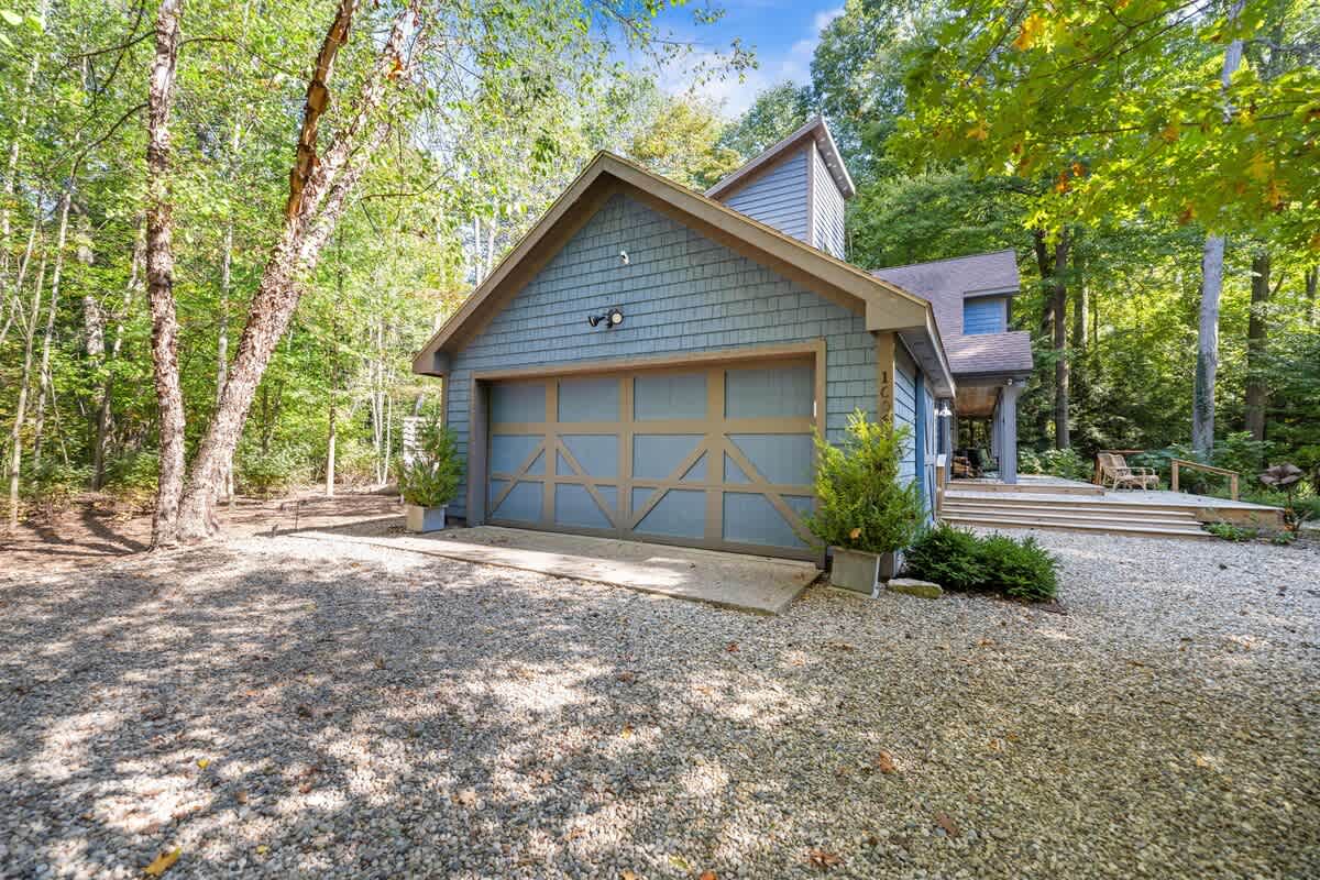 Front exterior view of a blue-shingled garage with a distinctive wooden garage door featuring a cross-brace design. The structure is nestled among lush green trees, with a gravel driveway leading up to it.